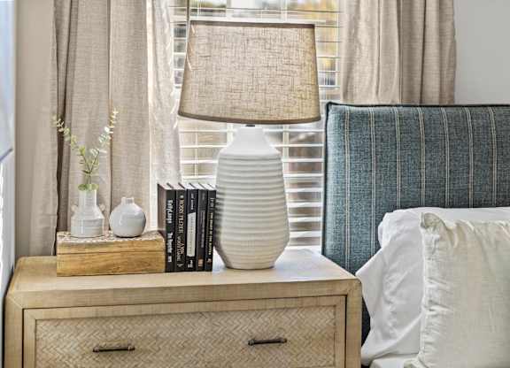 A lamp with a beige shade sits on a wooden nightstand next to a blue striped pillow at West Oak Apartments, North Carolina