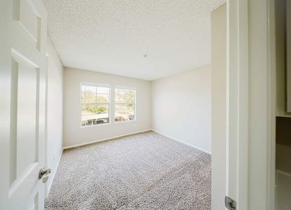 A room with a carpeted floor and a window with a view of trees and a balcony. at Legends at Rancho Belago, Moreno Valley, CA, 92553