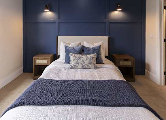 A bed with a white headboard and a blue blanket is in the center of the image at The Dean at Creekview Apartments, Newark