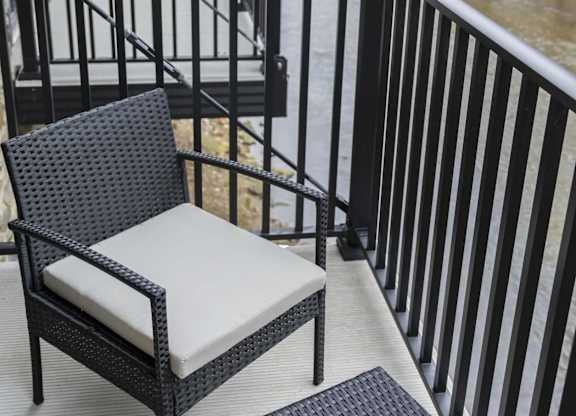 A black wicker chair with a white cushion sits on a balcony overlooking a river at The Dean at Creekview Apartments, Newark, Delaware