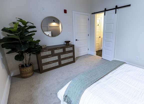 A bedroom with a bed, dresser, mirror, and plant at The Dean at Creekview Apartments, Newark, DE