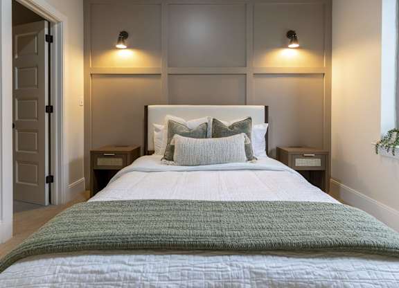 A bed with a green blanket and white pillows in a room with two wall lamps at The Dean at Creekview Apartments, Newark, DE