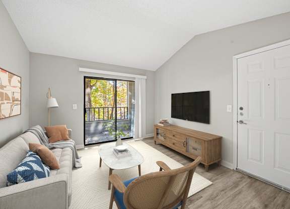 A living room with a grey couch, a white coffee table, a wooden chair, a flat screen TV, and a sliding glass door at Brampton Moors, Cary, NC, 27513