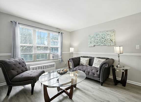 A living room with a grey couch, a black chair, and a glass coffee table at Huntwood Courts Apartments, Washington DC