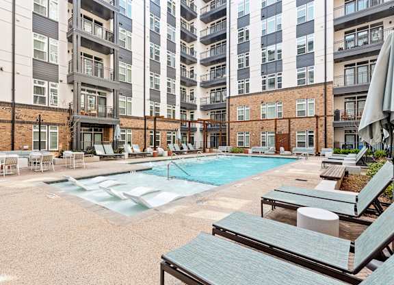 A large outdoor pool area with sun loungers and umbrellas in front of apartment buildings at Hawkins Press Apartments, North Carolina, 28203