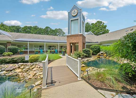 A small building with a clock tower is surrounded by a garden and a bridge.