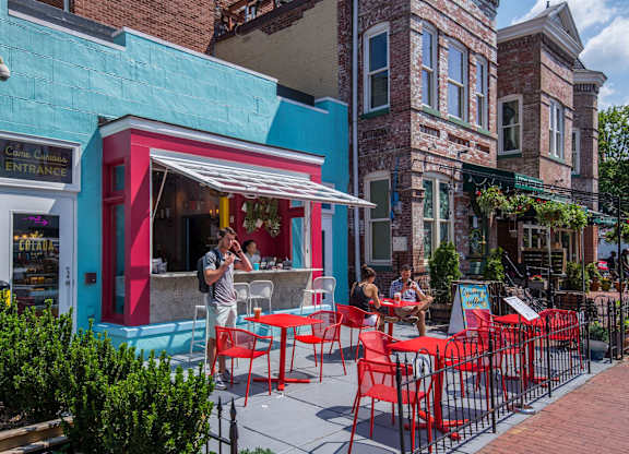A man is standing outside a restaurant with red chairs and tables.