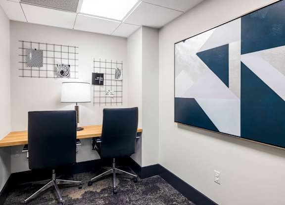 Two black chairs are placed in front of a desk with a white lamp and a painting on the wall.