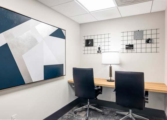 Two black chairs are in front of a desk with a lamp and a painting on the wall.