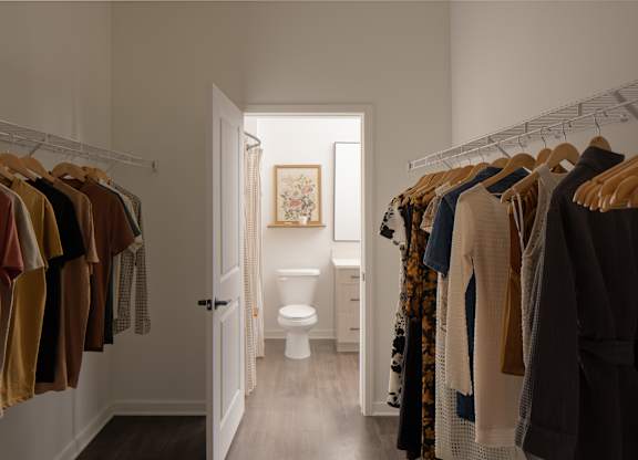 A hallway with clothes hanging on both sides and a toilet in the background.