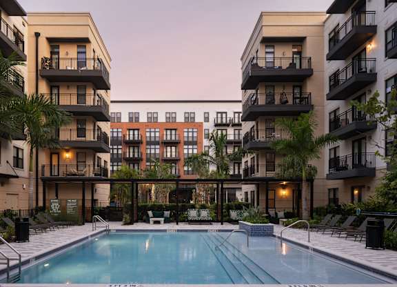 A swimming pool is surrounded by lounge chairs and palm trees in front of apartment buildings.