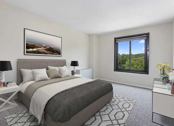 A bedroom with a large bed, a nightstand with a lamp, a framed picture on the wall, and a window with a view of trees.