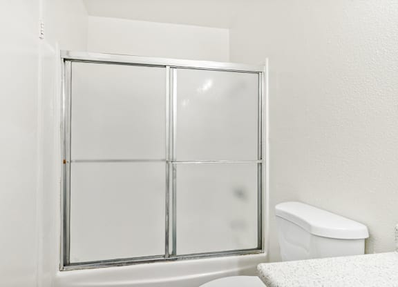 A white toilet sits next to a glass shower door in a bathroom. at Canyon Shadows Apartments, Riverside, CA 92503
