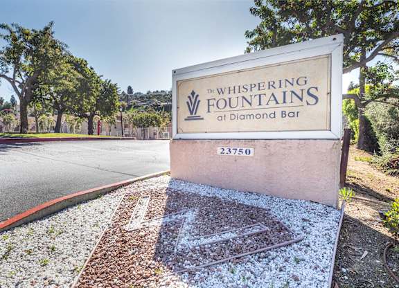 A sign for Whispering Fountains at Diamond Bar stands on a gravel area. at Whispering Fountains Diamond Bar, Diamond Bar, 91765
