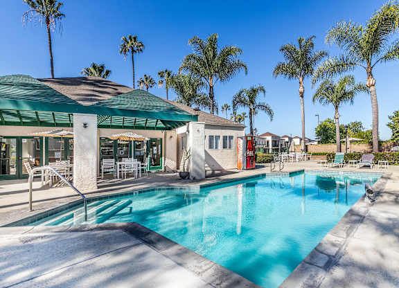 A pool surrounded by palm trees and a building with a green roof. at Whispering Fountains Lakewood, Lakewood Bar, CA