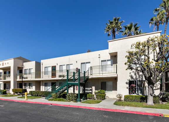 A white apartment building with green staircases and trees in front. at Whispering Fountains Lakewood, Lakewood Bar California