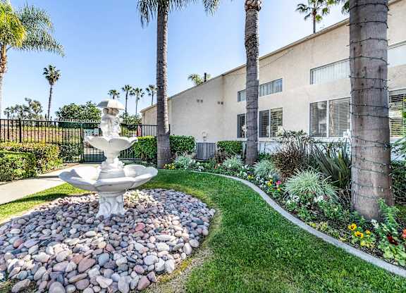A white fountain in the middle of a garden with a building in the background. at Whispering Fountains Lakewood, Lakewood Bar California