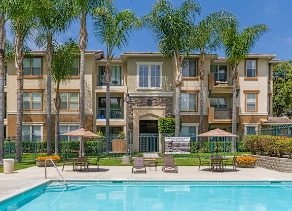 A swimming pool is surrounded by lounge chairs and umbrellas in front of a large apartment building.