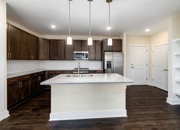 Open kitchen area with brown cabinets, stainless steel appliances and a sink in the island. book shelves off to the side.