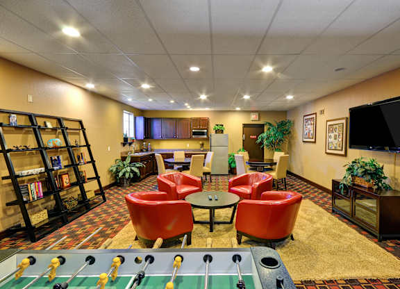 A foosball table is in the middle of a room with red chairs and a bookshelf.