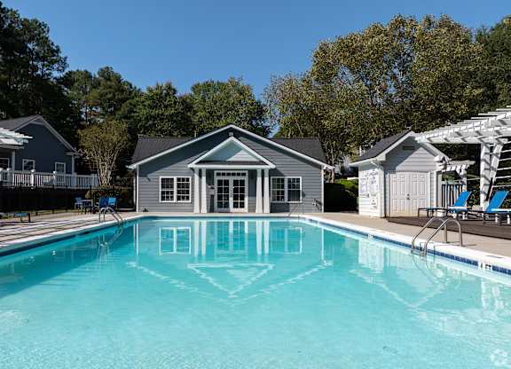 A swimming pool in front of a house with a deck and lounge chairs.