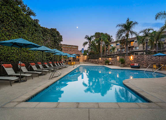 A pool surrounded by lounge chairs and umbrellas at dusk.