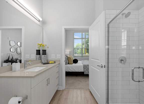A white bathroom with a walk-in shower and a bed in the background.