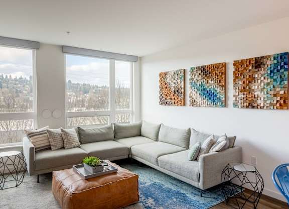 A living room with a grey couch, a brown leather ottoman, and three abstract paintings on the wall.at Axle, Washington, 98119