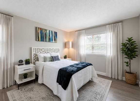 A bedroom with a large bed, a nightstand, a plant, and a painting on the wall.