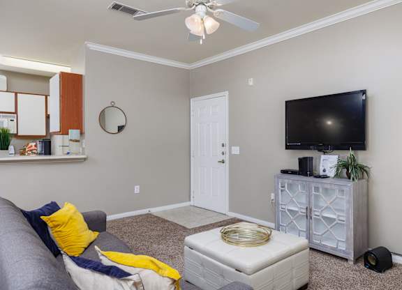 A living room with a grey couch, a white ottoman, a flat screen TV, and a ceiling fan at Clear Creek Meadows Apartments, Texas