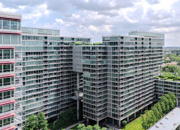 A modern glass building with a green roof is surrounded by trees and other buildings.