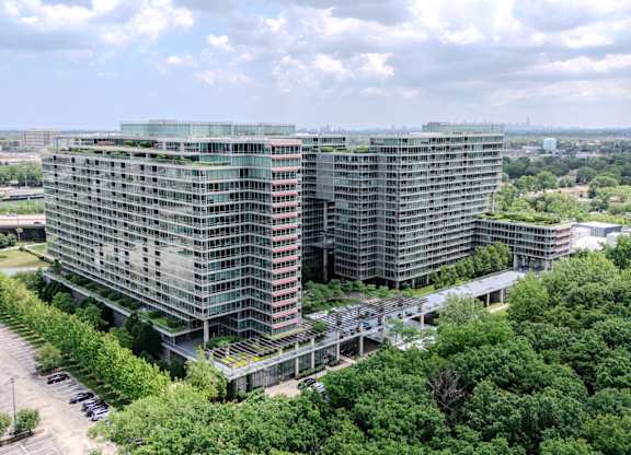A large, modern building with a glass facade is surrounded by a parking lot and greenery.