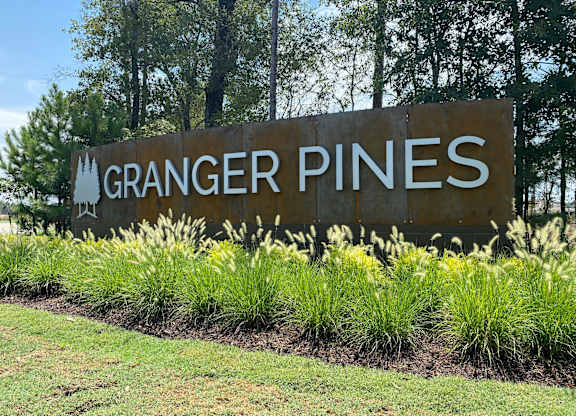 A sign that says "Granger Pines" is surrounded by grass and plants.
