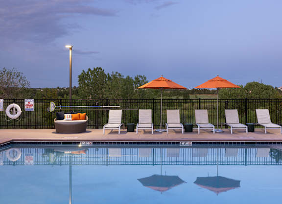 A pool with chairs and umbrellas is surrounded by a fence.