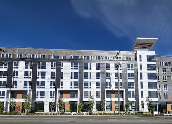 A large, modern multi-story building with a flat roof and a mix of white and dark grey balconies.