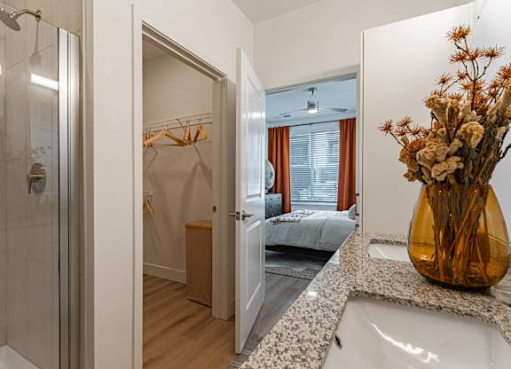 A bathroom with a walk-in shower, a bedroom, and a vase of dried flowers on the counter.