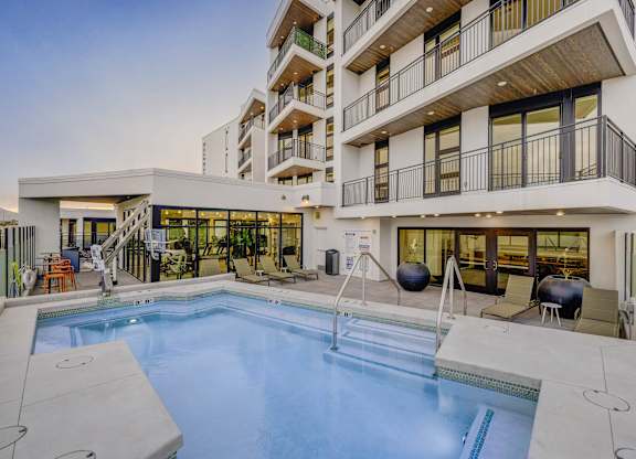 A large swimming pool is surrounded by a patio area with chairs and a hot tub.