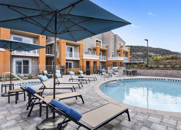 A pool area with sun loungers and umbrellas in front of apartment buildings.