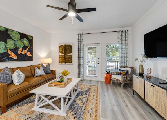 Charleston, SC, Apartments with In-Unit Washer and Dryer - The Avenues at Verdier Pointe - Living Room with Wood-Style Flooring, Ceiling Fan with Light, Couch, Armchair, Coffee Table, Painting, Rug, Media Console, and a Mounted TV.