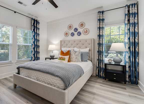 Dog-Friendly Apartments in Charleston, SC - The Avenues at Verdier Pointe - Bedroom with Wood-Style Flooring, Ceiling Fan, Bed, Nightstands, Lamps, and Large Windows with Blinds and Curtains.