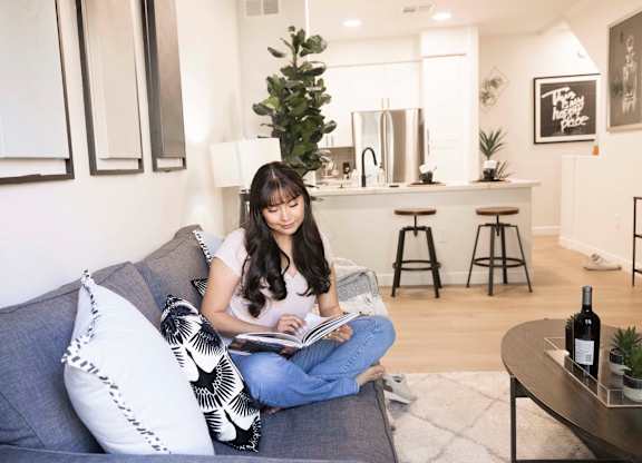 A woman is sitting on a grey couch reading a book.