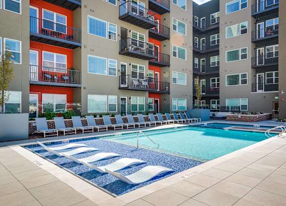 A swimming pool surrounded by lounge chairs in front of apartment buildings.