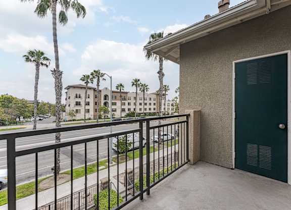 A balcony with a green door and a black railing overlooks a street with palm trees.