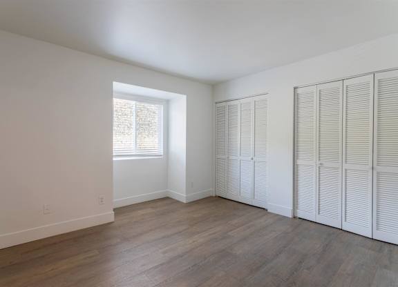 A room with white walls and wooden floors, with a window on the left and a set of white closet doors on the right.