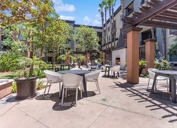 A patio with tables and chairs is surrounded by plants and trees at The Kitt at Warner Center Apartments, Woodland Hills 91303