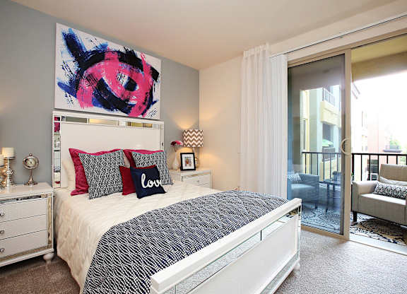 A bedroom with a large bed, a painting above it, and a chair in the doorway at The Kitt at Warner Center Apartments, Woodland Hills, CA