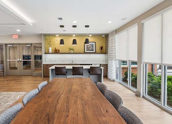 A long wooden table is surrounded by grey chairs in a room with a kitchen in the background.