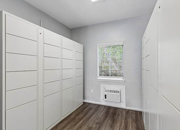 A room with white walls and wooden floors, featuring a row of white cupboards and a window with blinds.