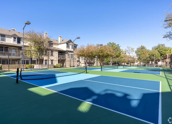 A tennis court with a blue surface and white lines is surrounded by apartment buildings.