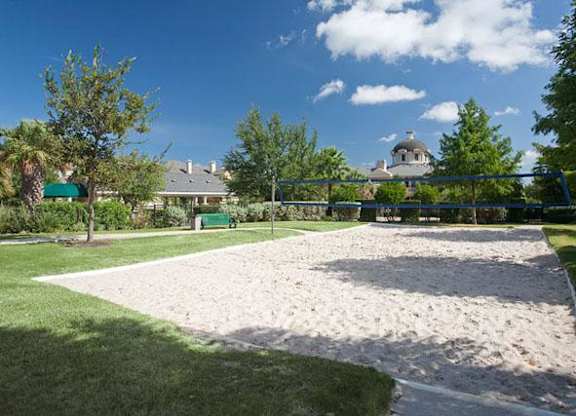 A sand pit in a park with a green fence and trees in the background.